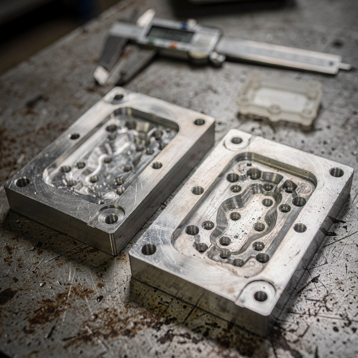 Two machined aluminum injection mold halves with a molded plastic part and calipers on a workbench
