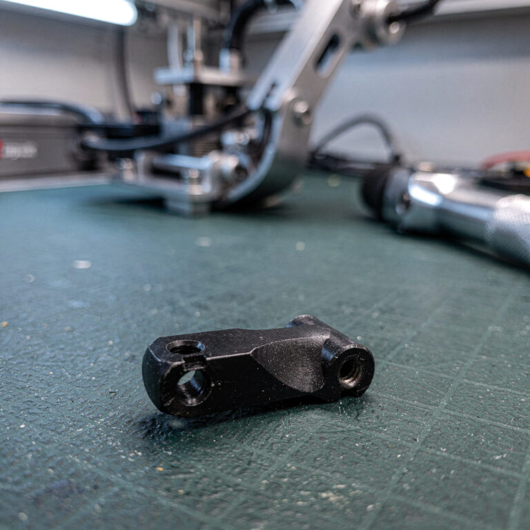 Black anodized aluminum robotic joint bracket on an ESD workbench mat with a robotic arm in the background
