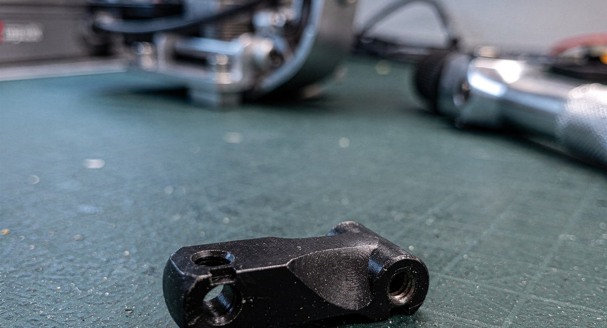 Black anodized aluminum robotic joint bracket on an ESD workbench mat with a robotic arm in the background
